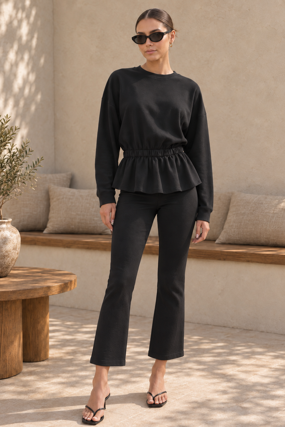 A woman modeling a black long-sleeve peplum top and matching black cropped flare pants, styled with black sunglasses and black heeled sandals in a sunlit neutral-toned indoor patio setting.