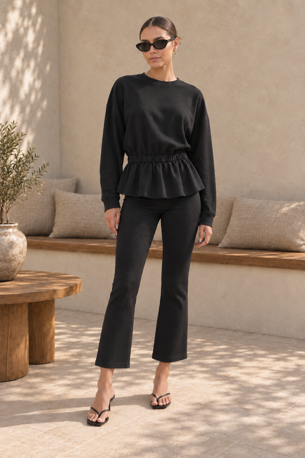 A woman modeling a black long-sleeve peplum top and matching black cropped flare pants, styled with black sunglasses and black heeled sandals in a sunlit neutral-toned indoor patio setting.