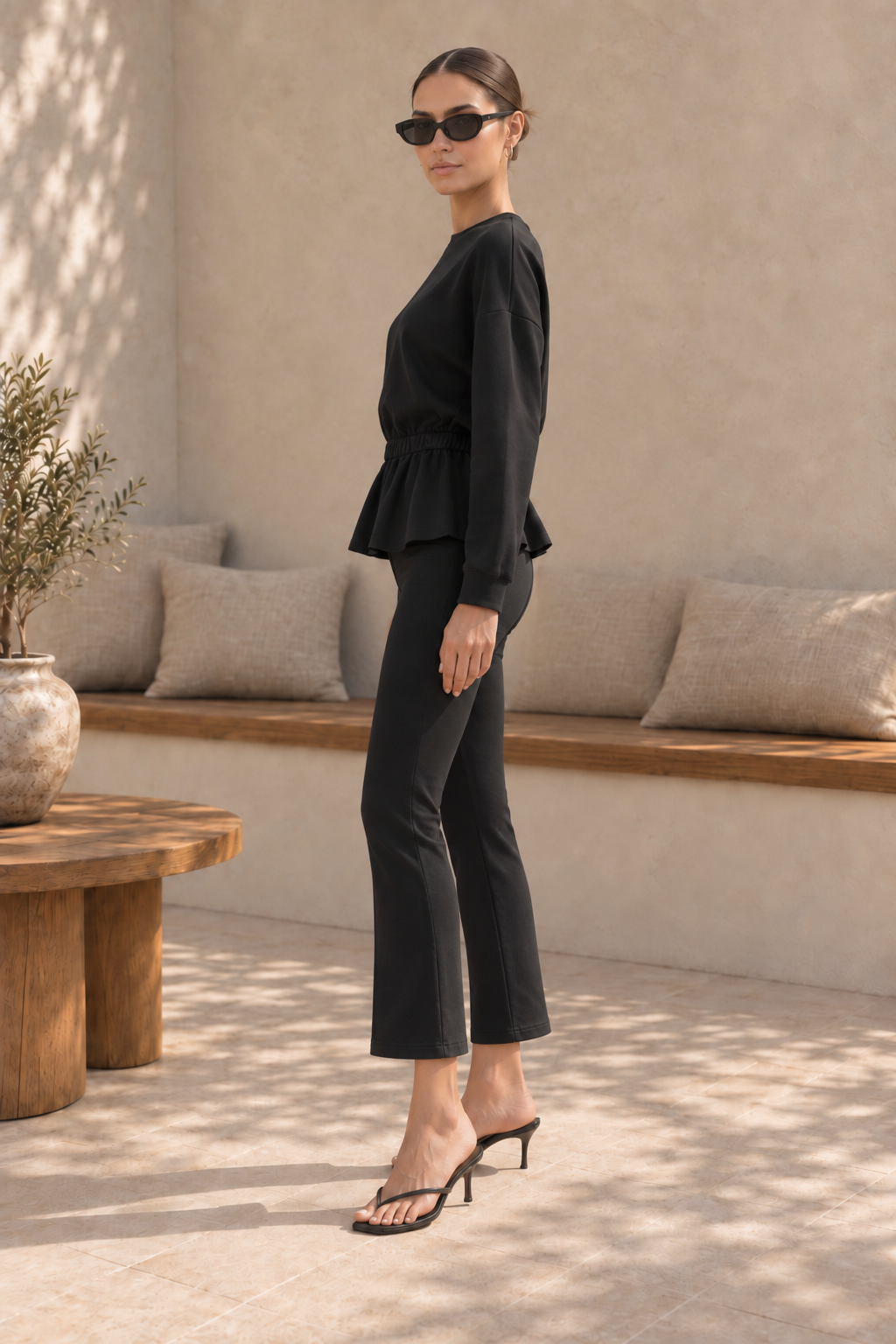 A woman modeling a black two-piece outfit consisting of a long-sleeve peplum top and matching cropped pants, styled with black sunglasses and black heeled sandals in a sunlit neutral-toned interior setting.