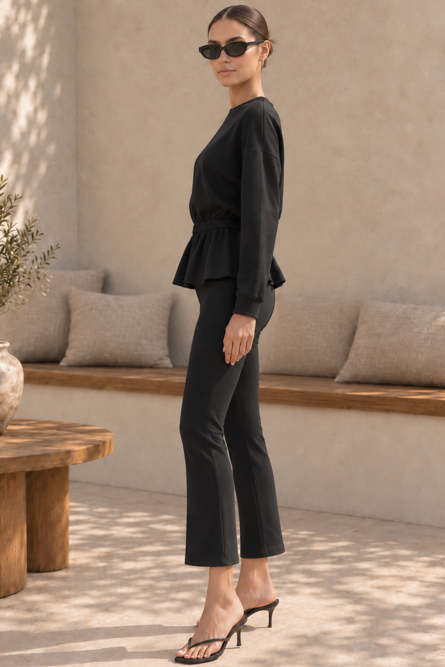 A woman modeling a black two-piece outfit consisting of a long-sleeve peplum top and matching cropped pants, styled with black sunglasses and black heeled sandals in a sunlit neutral-toned interior setting.