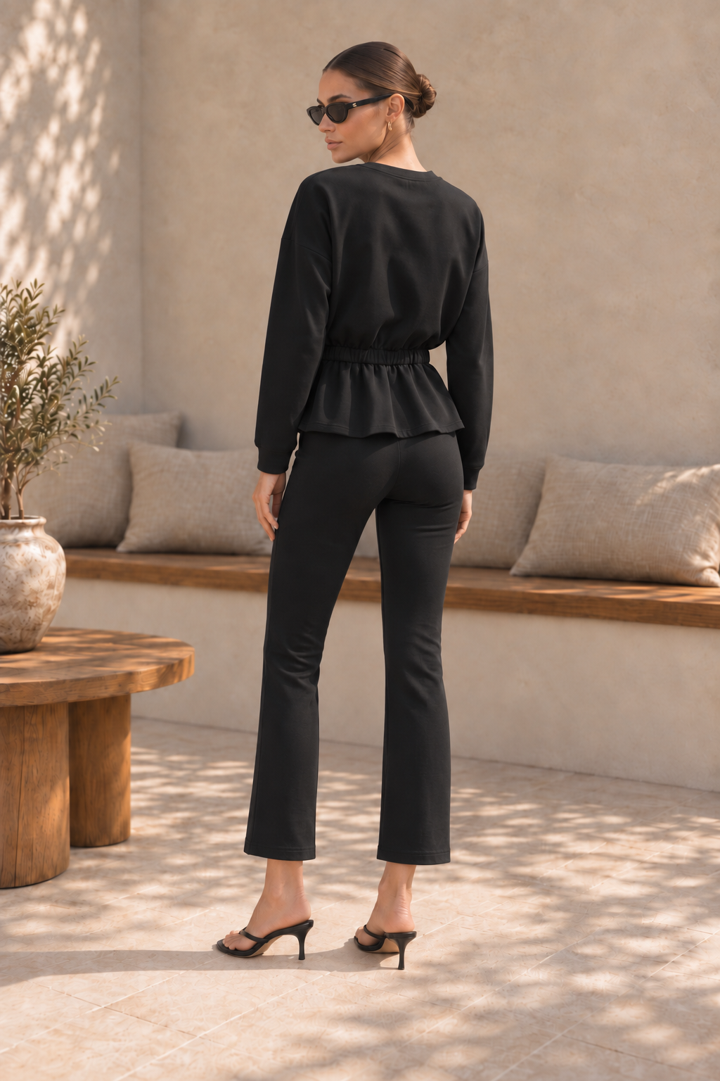 A woman stands with her back to the camera wearing a black long-sleeve peplum top and matching black cropped pants, styled with black heeled sandals in a sunlit patio setting.