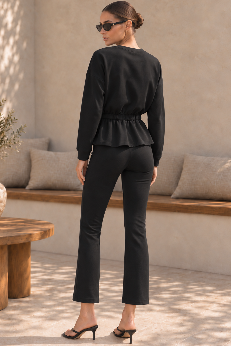 A woman stands with her back to the camera wearing a black long-sleeve peplum top and matching black cropped pants, styled with black heeled sandals in a sunlit patio setting.