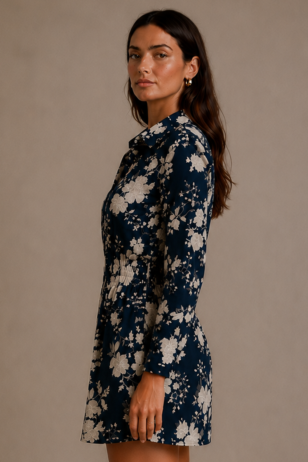 A woman modeling a navy blue long-sleeve mini dress with a white floral pattern, photographed against a solid warm beige studio background.