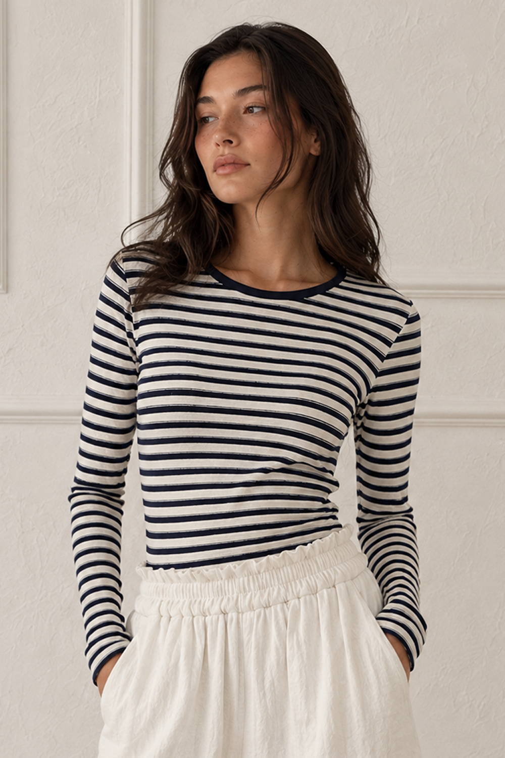 A woman wearing a navy and white horizontally striped long-sleeve top with a crew neck, paired with an off-white skirt, standing against a white paneled wall.