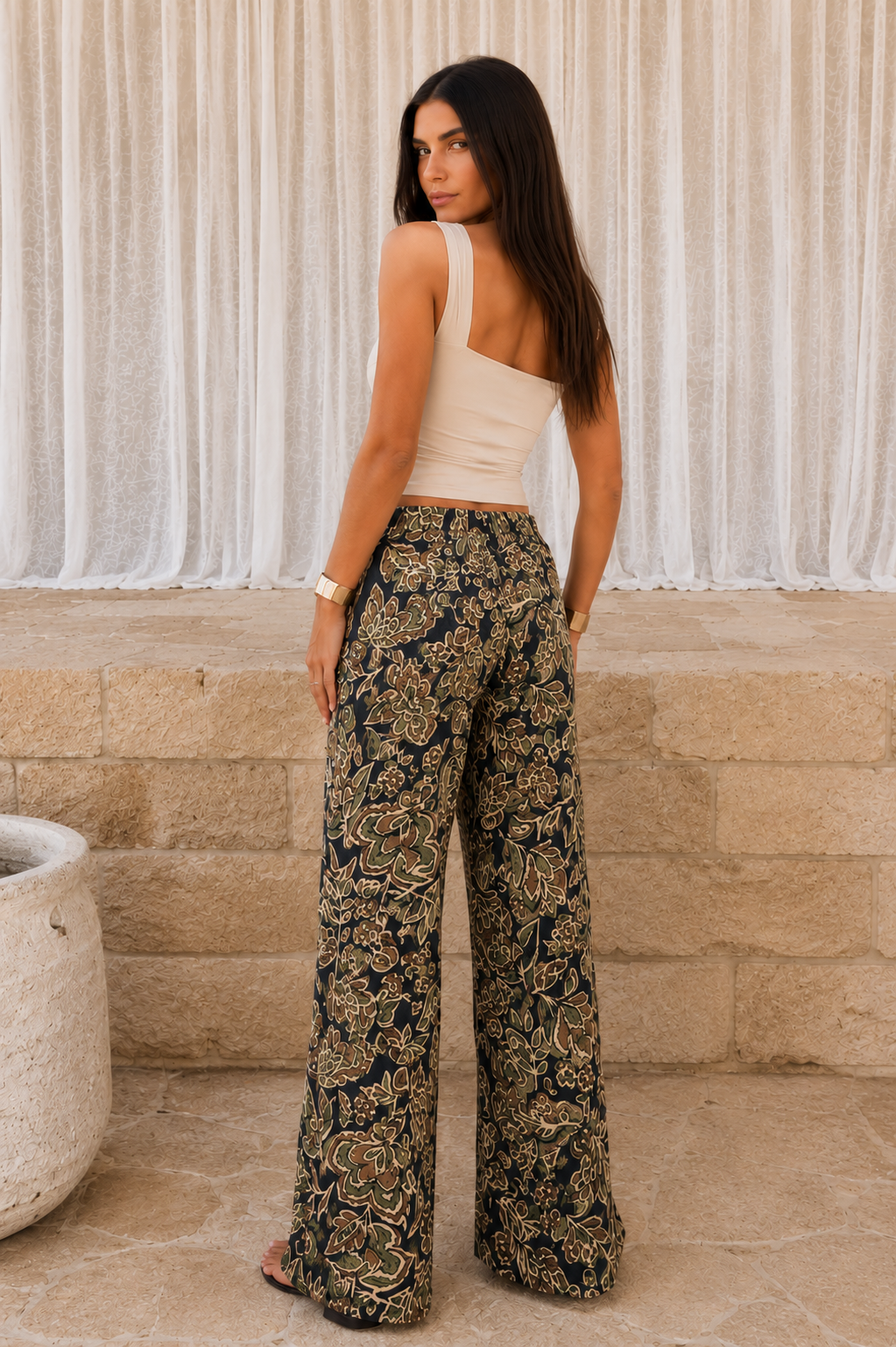 Woman wearing a beige top and floral pants standing against a textured wall.