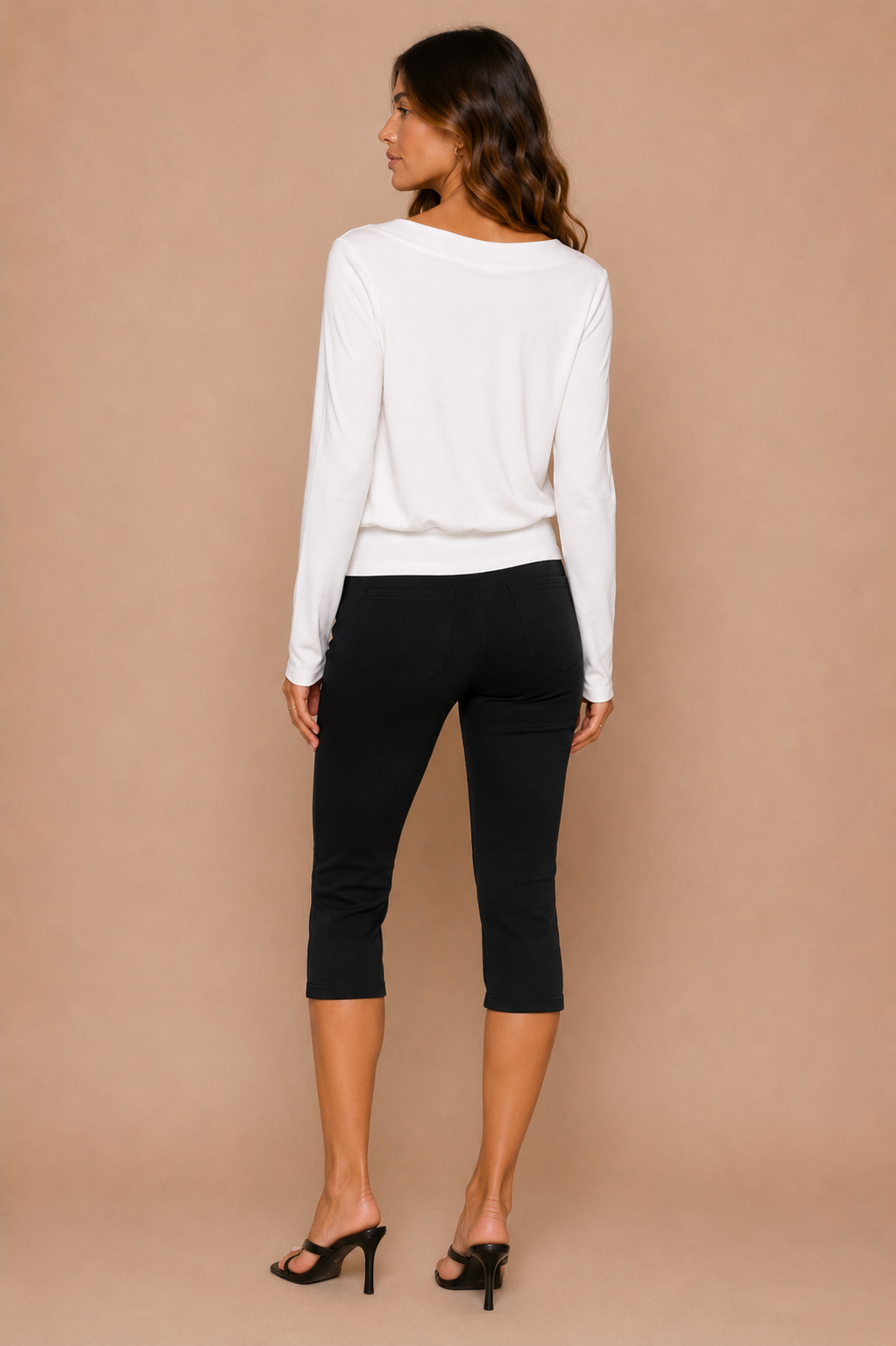 A brunette woman wearing white top and black capri pants in front of beige backdrop.