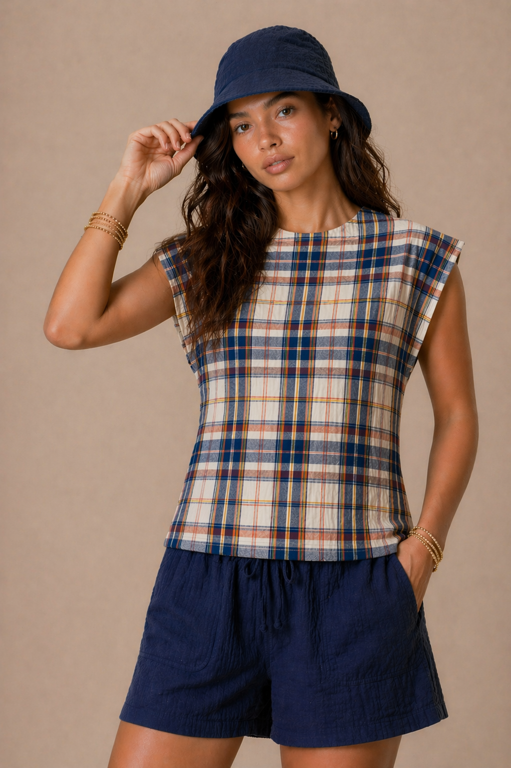 Woman wearing a plaid top and navy shorts with a navy bucket hat against a beige background