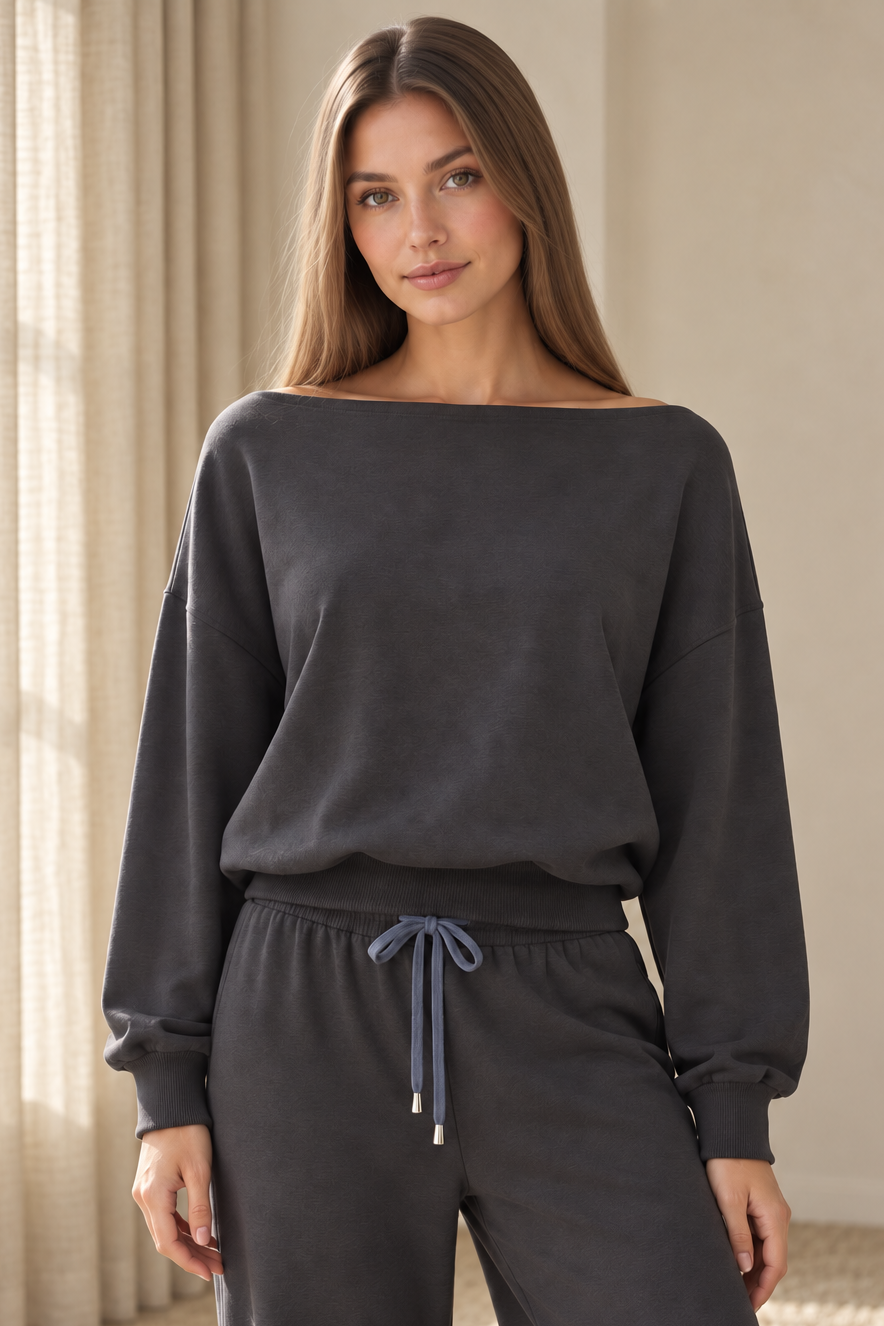 A woman wearing a dark charcoal gray off-the-shoulder sweatshirt and matching sweatpants with a contrasting blue-gray drawstring, standing in a softly lit indoor setting.