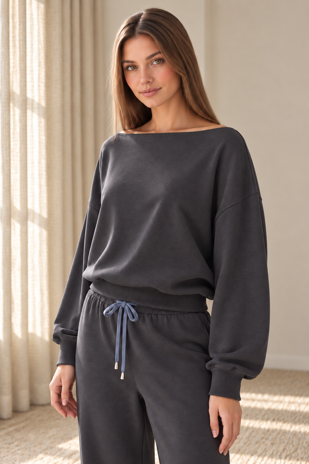 A woman modeling a dark charcoal gray lounge set consisting of an off-the-shoulder long-sleeve sweatshirt and matching drawstring sweatpants in a brightly lit room with beige curtains.