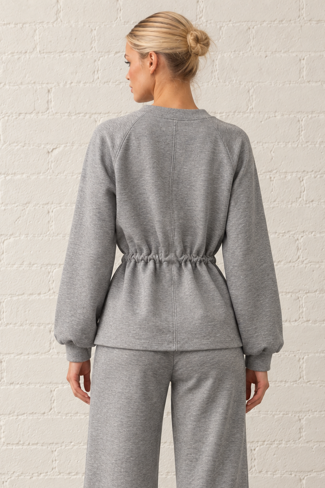 A woman viewed from behind wearing a heather gray two-piece lounge set consisting of a long-sleeve cropped sweatshirt with a drawstring waist and matching wide-leg sweatpants, standing against a white brick wall.