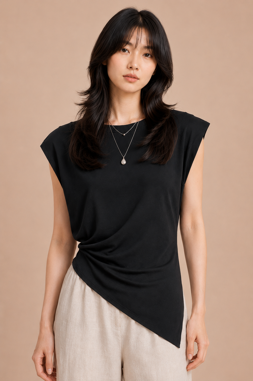 Woman wearing black asymmetrical top and linen pants against beige backdrop.