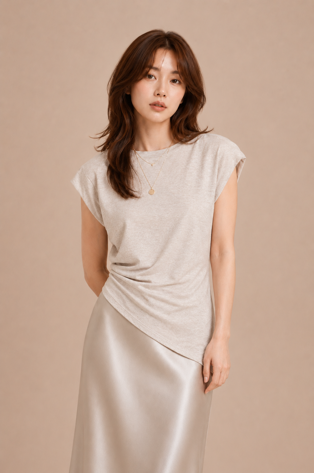 Woman wearing beige asymmetrical top and silk skirt against beige backdrop.